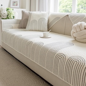 Puede incluir: Sofá color crema con un patrón geométrico de líneas curvas negras. Se ven dos cojines a juego y una funda de sofá con el mismo diseño. Una taza y un platillo blancos están sobre el sofá.