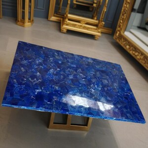 May include: Rectangular coffee table with a mosaic top of deep blue stones. The table has a gold-colored metal base. The tabletop is made of many small, irregularly shaped pieces of stone. The table is a statement piece.