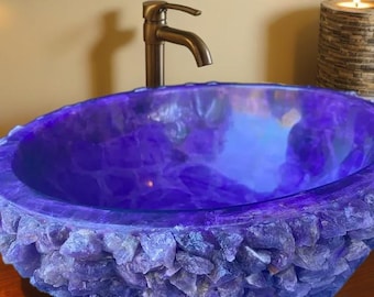 May include: A striking, handcrafted amethyst sink with a rough, crystalline exterior and a smooth, polished interior. The sink is a deep purple color and features a bronze faucet. A candle in a decorative holder is visible in the background.