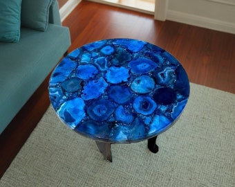 May include: A round blue agate table with a polished finish. The table has a unique design with a variety of blue agate slices. The table is supported by three black legs.