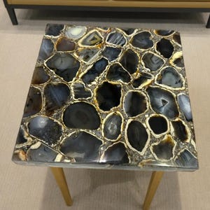 May include: Square coffee table with a mosaic top made of polished agate stones in shades of black, gray, and cream. The stones are set in a light-colored resin and framed by a thin, light-colored border. The table has four slender, light-colored wooden legs.