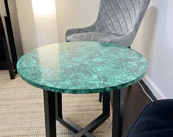 Puede incluir: Una mesa redonda verde con una base de metal negro y un soporte en forma de cruz. La mesa está hecha de malaquita, una piedra preciosa verde. Se puede ver una silla gris en el fondo.