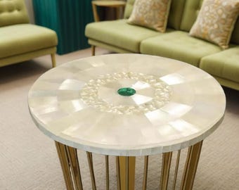 Puede incluir: Una mesa auxiliar redonda con una tapa con incrustaciones de nácar y patas de metal dorado. La parte superior de la mesa presenta una gema verde central y un patrón decorativo. Es un mueble decorativo.