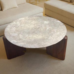 Pode incluir: Uma mesa de centro redonda com um tampo em estilo mosaico em tons de branco e cinza. A mesa tem uma base de madeira maciça castanho escuro. Uma peça de mobiliário moderna.