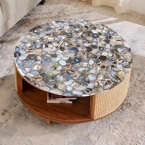 May include: Round coffee table with a mosaic stone top in shades of gray, blue, and white. The table has a wooden base with a lower shelf and a woven rattan side panel. The table is approximately 36 inches in diameter.