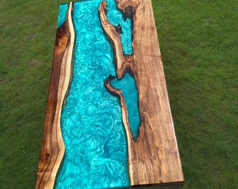 May include: A wooden table with a turquoise epoxy resin river flowing through the center. The table has a natural wood finish and black metal legs.