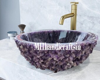 May include: A purple amethyst stone sink with a gold faucet. The sink is made of many small amethyst crystals and has a smooth, polished interior. The faucet is tall and slender with a square base. The sink is sitting on a white countertop.