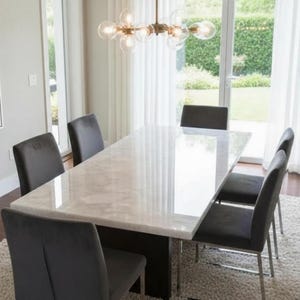 Puede incluir: Comedor moderno con una mesa rectangular con tapa de mármol y seis sillas grises tapizadas. Una lámpara de araña dorada cuelga sobre la mesa. La habitación tiene una gran ventana con cortinas blancas y una pintura. La mesa mide aproximadamente 244 cm de largo.