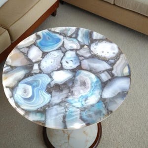 May include: Round coffee table with a mosaic top made of blue, white, and gray agate stones. The table has a white marble-like base and a dark brown wooden trim. The table is a decorative furniture piece.
