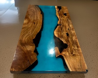 May include: A handcrafted wooden serving board with a vibrant blue epoxy resin river. The board features natural wood grain patterns and a glossy finish. The board is approximately 18 inches long and 12 inches wide.