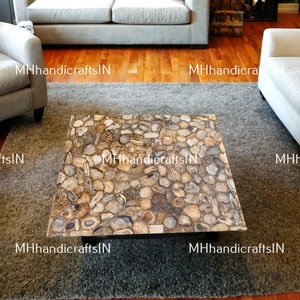 Puede incluir: Una mesa de centro cuadrada con una tapa hecha de rodajas de ágata en tonos marrones, beige y grises. La mesa está sobre una alfombra gris en un salón.