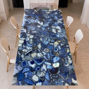 Könnte beinhalten: Ein rechteckiger Esstisch mit einer blauen Achat-Tischplatte und goldenen Metallbeinen. Der Tisch ist von vier weißen Stühlen mit goldenen Metallbeinen umgeben.