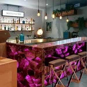 Può includere: Un bar con un piano di lavoro viola illuminato e sgabelli abbinati. Il bar ha un'insegna con la scritta "SCOTCH & TED" ed è illuminato da lampade a sospensione. Scaffali dietro il bar espongono bottiglie.