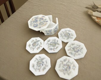 May include: Set of seven white octagonal coasters with floral designs in blue and silver. The coasters are made of marble and have a raised edge. The coasters are arranged in a circle on a beige tablecloth.