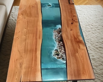 May include: A rectangular coffee table with a unique design. The table features two wooden planks on either side of a central section filled with blue resin, resembling water. The resin section includes small stones, creating a coastal theme. The table is supported by black metal legs.