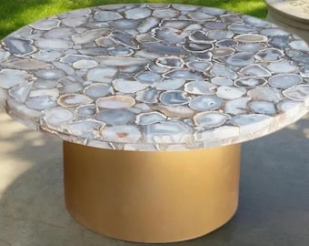 May include: A round coffee table with a mosaic top made of gray and white agate stones. The table has a cylindrical, gold-colored base. This unique table is a statement piece for any living room.