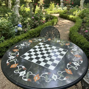 Puede incluir: Mesa redonda de mármol verde oscuro con tablero de ajedrez incorporado e incrustaciones florales decorativas. La mesa está en un jardín exuberante con estatuas y un camino sinuoso. Diámetro aproximado de 122 cm.