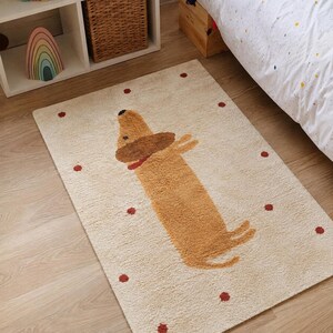 May include: A rectangular area rug in a cream color featuring a central graphic of a tan dachshund dog with a brown head and a red collar. The rug is decorated with small, scattered red circular dots across the entire surface of the fabric.