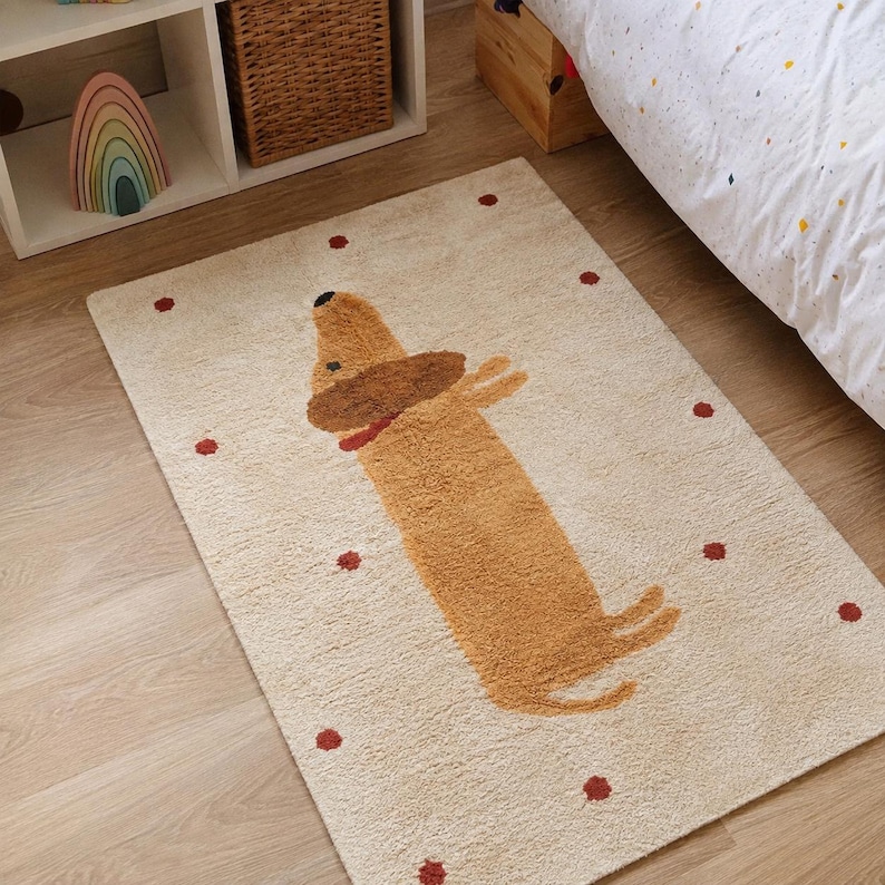 May include: A rectangular area rug in a cream color featuring a central graphic of a tan dachshund dog with a brown head and a red collar. The rug is decorated with small, scattered red circular dots across the entire surface of the fabric.