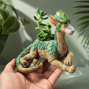 Peut inclure: Un pot de fleurs décoratif en forme de créature mythique avec un corps brun texturé rappelant le grain du bois et des accents verts en forme de feuilles. La créature a des oreilles pointues, des yeux jaunes et une longue queue, avec une plante succulente verte poussant sur son dos.