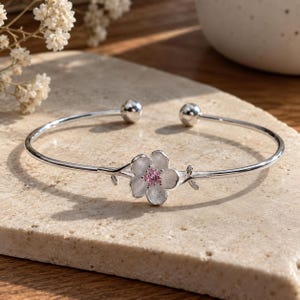 Peut inclure: Un bracelet manchette en argent arborant une fleur à cinq pétales en son centre, ornée d'une petite pierre précieuse rose ronde. Le bracelet présente une finition polie, un anneau fin et deux petites terminaisons sphériques aux extrémités.