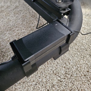 May include: A black rectangular device with a textured top is attached to a curved black metal tube. The device is secured with black clips and has a power cord connected. The background is a light-colored carpet.