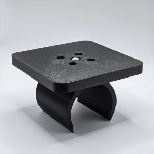 May include: A small, black, square-shaped table with a curved base. The tabletop has four small holes and a silver-colored fastener in the center. The table is made of a textured, matte material.