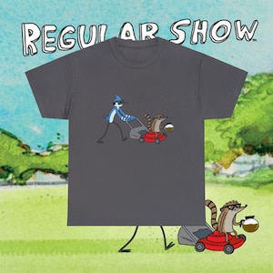 May include: Dark gray t-shirt featuring a cartoon image of Mordecai and Rigby from Regular Show mowing the lawn. The text "REGULAR SHOW" is above the graphic in a stylized font. The shirt is made of a soft material.