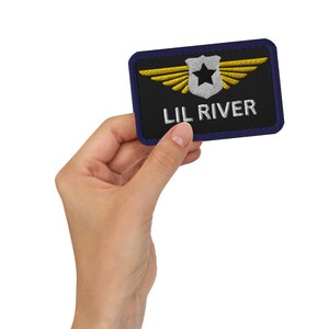 May include: A black embroidered patch with a gold winged shield and a white star. The text "LIL RIVER" is embroidered in white on the patch.