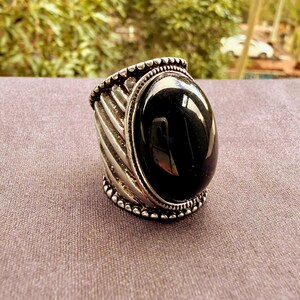 May include: A silver ring featuring a large, smooth, oval-shaped black stone set in the centre. The band is wide and decorated with diagonal, raised ridges and a beaded border along the top and bottom edges of the metal setting.