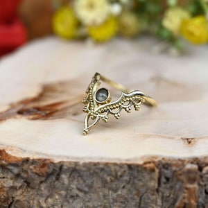 Può includere: Un anello color oro con un design a forma di corona. L'anello presenta una gemma grigia rotonda centrale e intricati dettagli in filigrana lungo la fascia. L'anello è appoggiato su una superficie di legno.