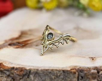 May include: A gold-colored ring with a crown-like design. The ring features a central, round, gray gemstone and intricate filigree detailing along the band. The ring is set on a wooden surface.
