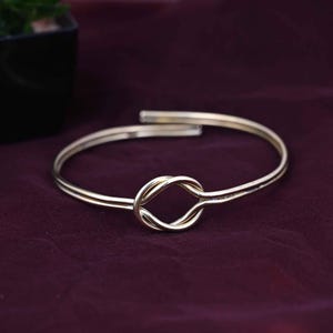 May include: A gold-colored bracelet with a knot design. The bracelet is made of thin, curved metal and features a knot in the center. The background is a dark purple color.
