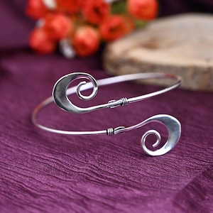 May include: A silver-toned arm cuff bracelet with a spiral design. The bracelet features two curved, open-ended sections with decorative swirls at each end. The cuff is displayed on a purple surface.