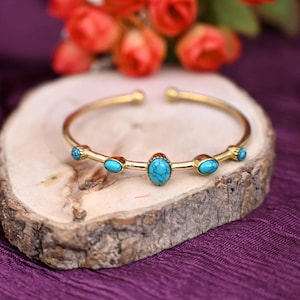 May include: A gold-colored cuff bracelet with turquoise-colored stones. The bracelet has an open design and is decorated with five oval-shaped turquoise stones. The bracelet is displayed on a wooden surface.