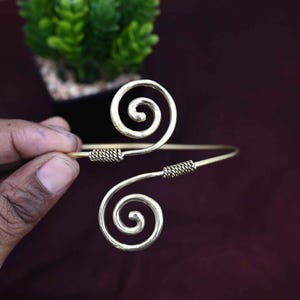 May include: A gold-colored arm cuff with a spiral design. The cuff features two large spirals connected by a straight bar. The bar has textured detailing near the spirals. The background is a dark maroon color.
