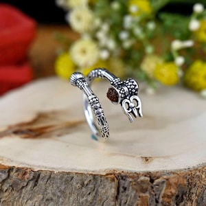 Può includere: Anello d'argento con un designo dettagliato che presenta un lingam Shiva e un tridente. L'anello è avvolto attorno a una pietra marrone.