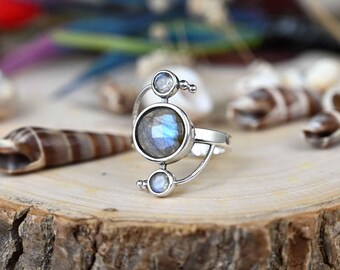 May include: A silver ring with a crescent moon design featuring three round labradorite gemstones. The ring is set on a wooden surface.