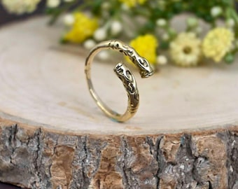 May include: A gold-colored adjustable ring with an open design. The ring features an engraved pattern on the two ends that meet. The ring is displayed on a wooden slice with yellow flowers in the background.