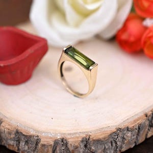 May include: A gold ring with a rectangular, light green gemstone. The ring is set on a wooden surface, with a red object and flowers in the background. The ring's band is a simple, rounded design.