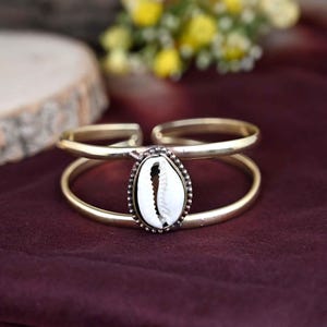 Może przedstawiać: Złota bransoletka z podwójną obręczą i białym zawieszką w kształcie muszli kauri.