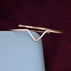 May include: A gold-colored metal bracelet with a V-shaped design. The bracelet is resting on a light blue surface, with a dark burgundy background. The bracelet's simple, geometric design makes it a versatile accessory.