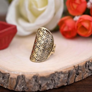 Pode incluir: Um anel dourado com um design vazado com um padrão geométrico. O anel está sobre uma superfície de madeira, com flores desfocadas ao fundo. O design do anel é uma série de formas interconectadas.