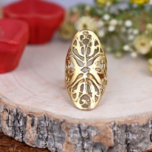 Peut inclure: Une bague en filigrane dorée de forme ovale avec un motif découpé complexe. La bague est posée sur une surface en bois.