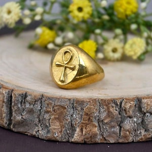 Puede incluir: Un anillo dorado con una cara ovalada que presenta un símbolo Ankh en relieve. El anillo está sobre una superficie de madera rústica, con flores amarillas y blancas en el fondo. El anillo tiene un acabado pulido.