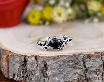 May include: A silver ring featuring a central black gemstone, with leaf-shaped embellishments on either side. The ring is displayed on a wooden surface, with blurred yellow and white flowers in the background. The ring's design is detailed.