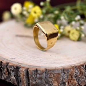 Könnte beinhalten: Ein goldfarbener Siegelring mit rechteckiger Oberfläche. Der Ring liegt auf einer Holzoberfläche mit einem unscharfen Hintergrund aus gelben und weißen Blumen. Der Ring hat ein glattes, abgerundetes Band.
