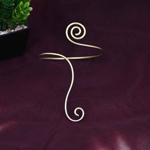 May include: A gold-colored arm cuff with a spiral design at the top and a curved, looping shape. The jewelry piece is set against a dark burgundy background, creating a contrast that highlights its form.