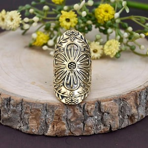 Könnte beinhalten: Ein goldfarbener, ovaler Ring mit einem aufwendigen Blumenmuster. Der Ring hat ein zentrales Blumenmotiv, umgeben von detaillierten Mustern, und liegt auf einem Holzuntergrund mit gelben Blumen.