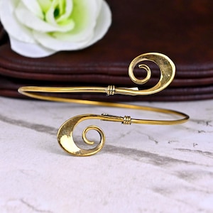 Puede incluir: Un brazalete de metal dorado con un diseño único en espiral. El brazalete es abierto y ajustable para adaptarse a diferentes tamaños de muñeca.